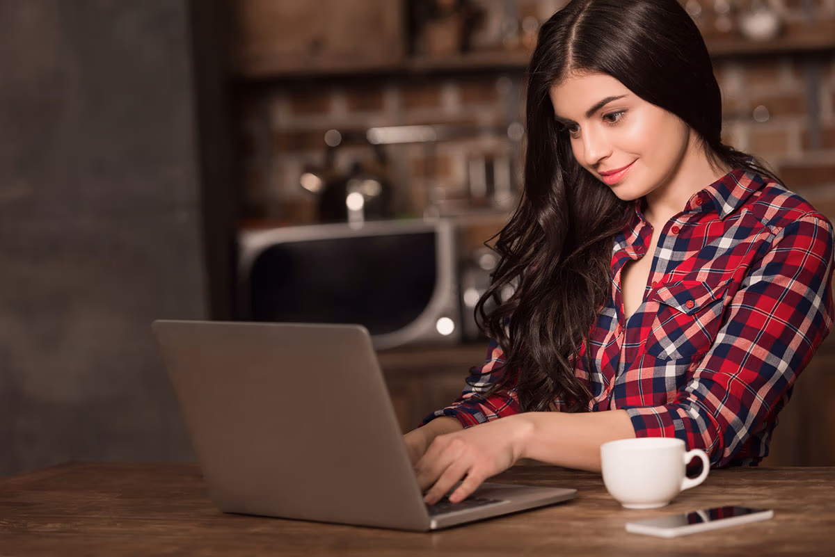 How to write a blog post: a girl typing on a laptop with a smartphone and coffee beside her.